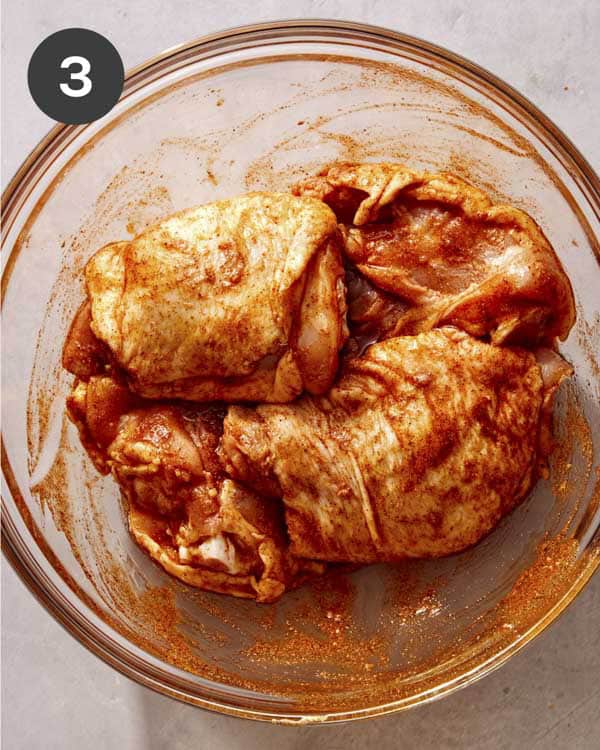 Chicken thighs tossed in seasoning in a glass bowl.