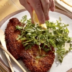 Chicken milanese on a dinner plate with someone squeezing lemon on it.
