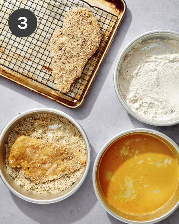 A chicken breast breaded on a baking sheet with the breading ingredients next to it.