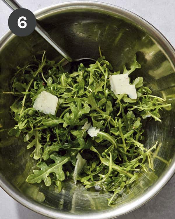 Arugula in a bowl with dressing and parmesan tossed together.