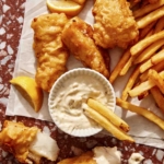 Beer battered fish on parchment with french fries and tartar sauce with some fries dipping in the sauce.