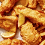 Close up on a bunch of beer battered fish on parchment with lemons and fries peeking in.