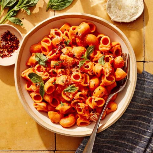 Spicy rigatoni in a bowl with some basil, red pepper flakes, and cheese on the side.