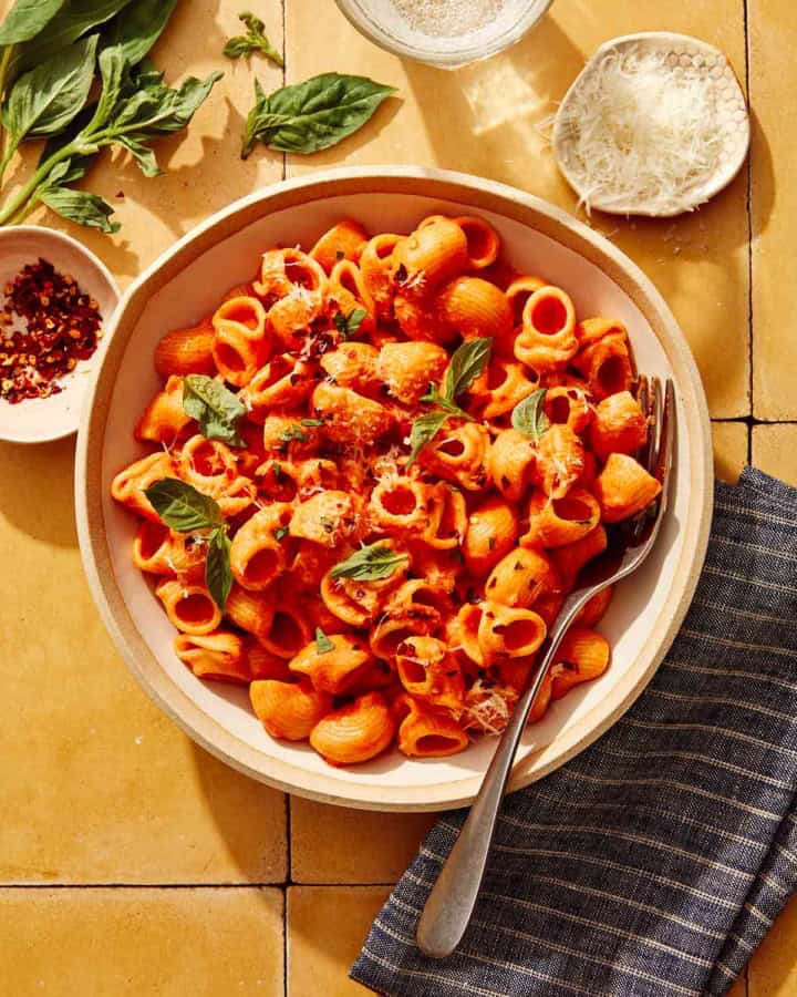 Spicy rigatoni in a bowl with some basil, red pepper flakes, and cheese on the side.