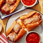 Two meatball subs on plates with marinara sauce on the side.