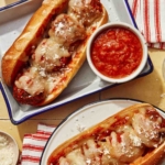 Two meatball subs on plates with marinara sauce on the side.