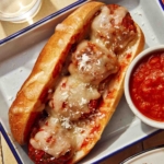 Close up on a meatball sub with water in the background and a side of marinara sauce.