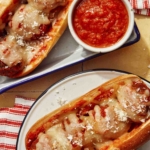 Two meatball subs on plates with marinara sauce on the side.