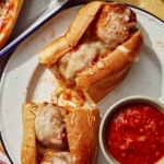 A meatball sub on a plate cut in half with marinara sauce on the side.