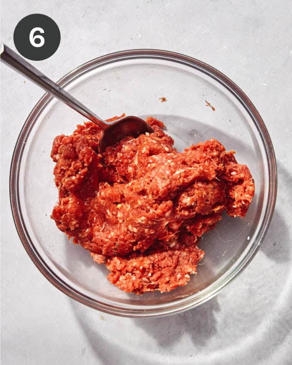 Ground beef in a glass bowl with a wooden spoon being mixed with spices.