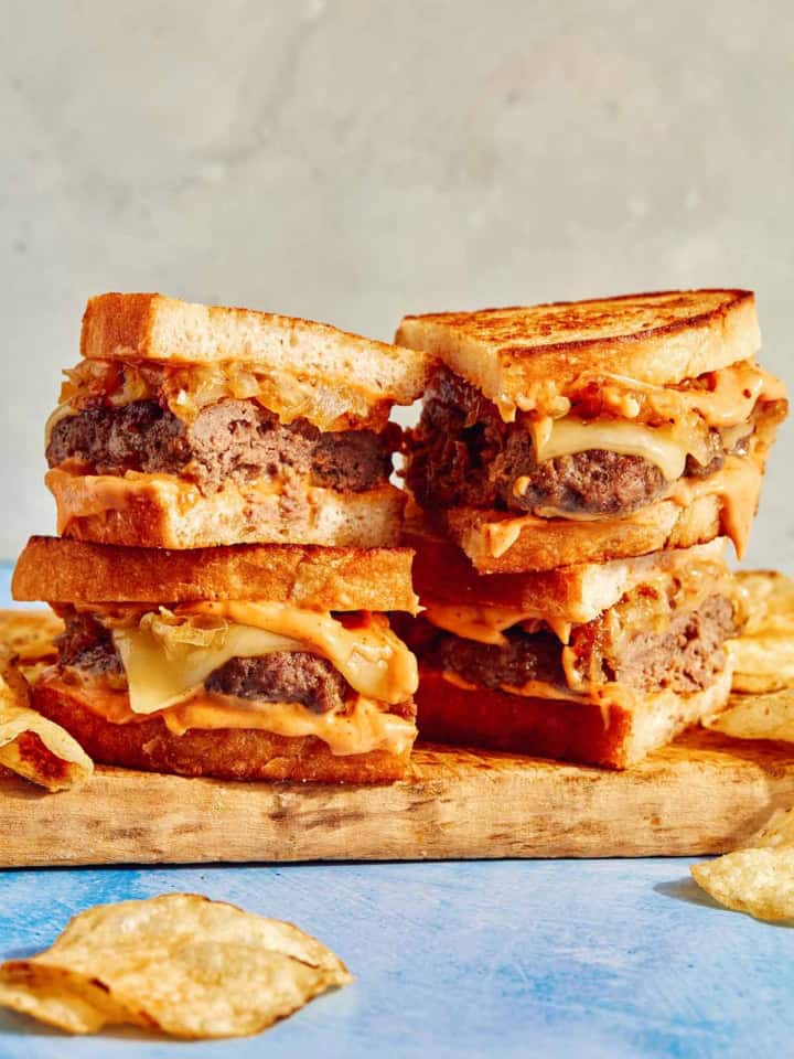 Two patty melts on a cutting board cut in half with chips on the side.