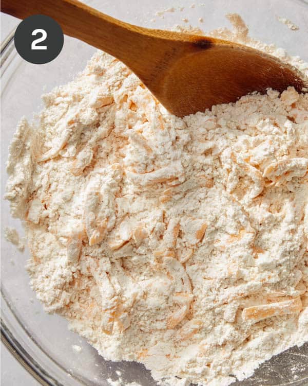 Flour mixture being mixed with shredded cheddar cheese in a bowl with a spoon.