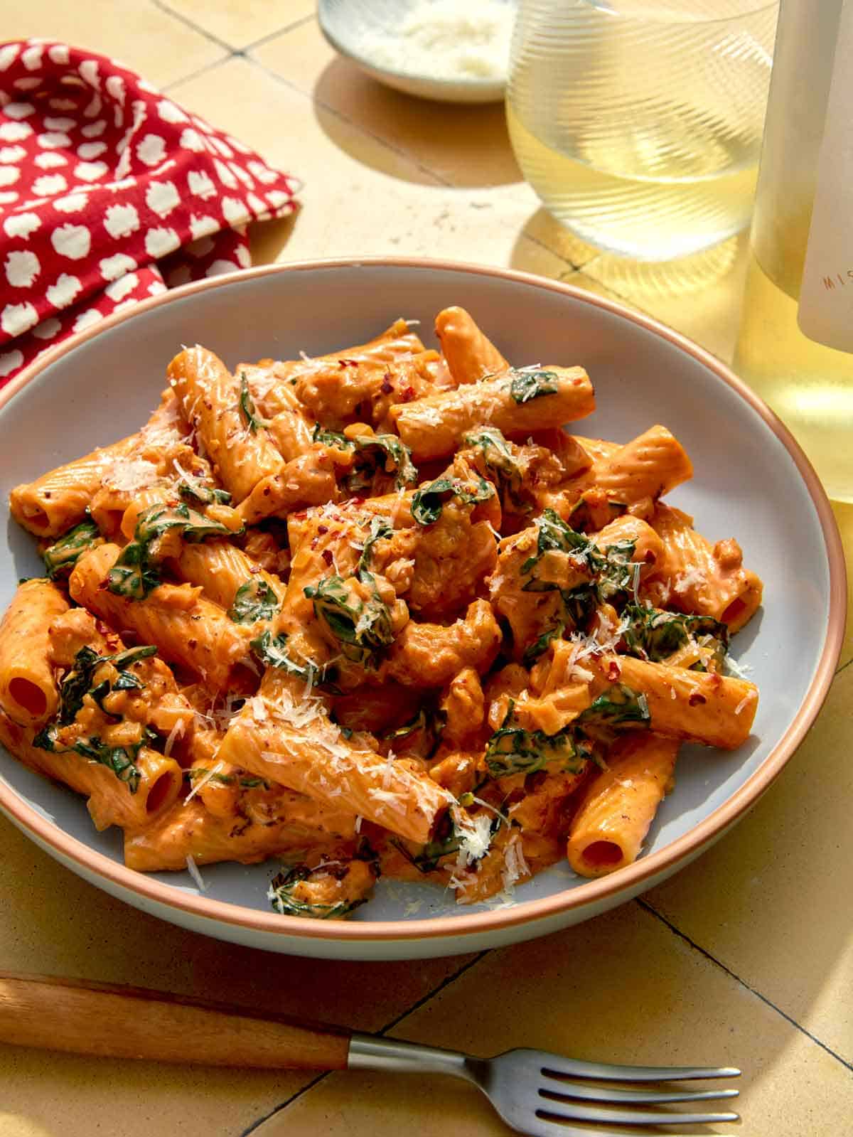 A dinner scene with a bowl of kale and sausage rigatoni with wine in the background.