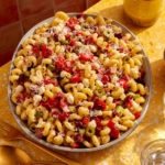 Pasta salad on a table with serving utensils and a jar of Italian vinaigrette.