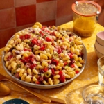 Pasta salad on a table with serving utensils and a jar of Italian vinaigrette.