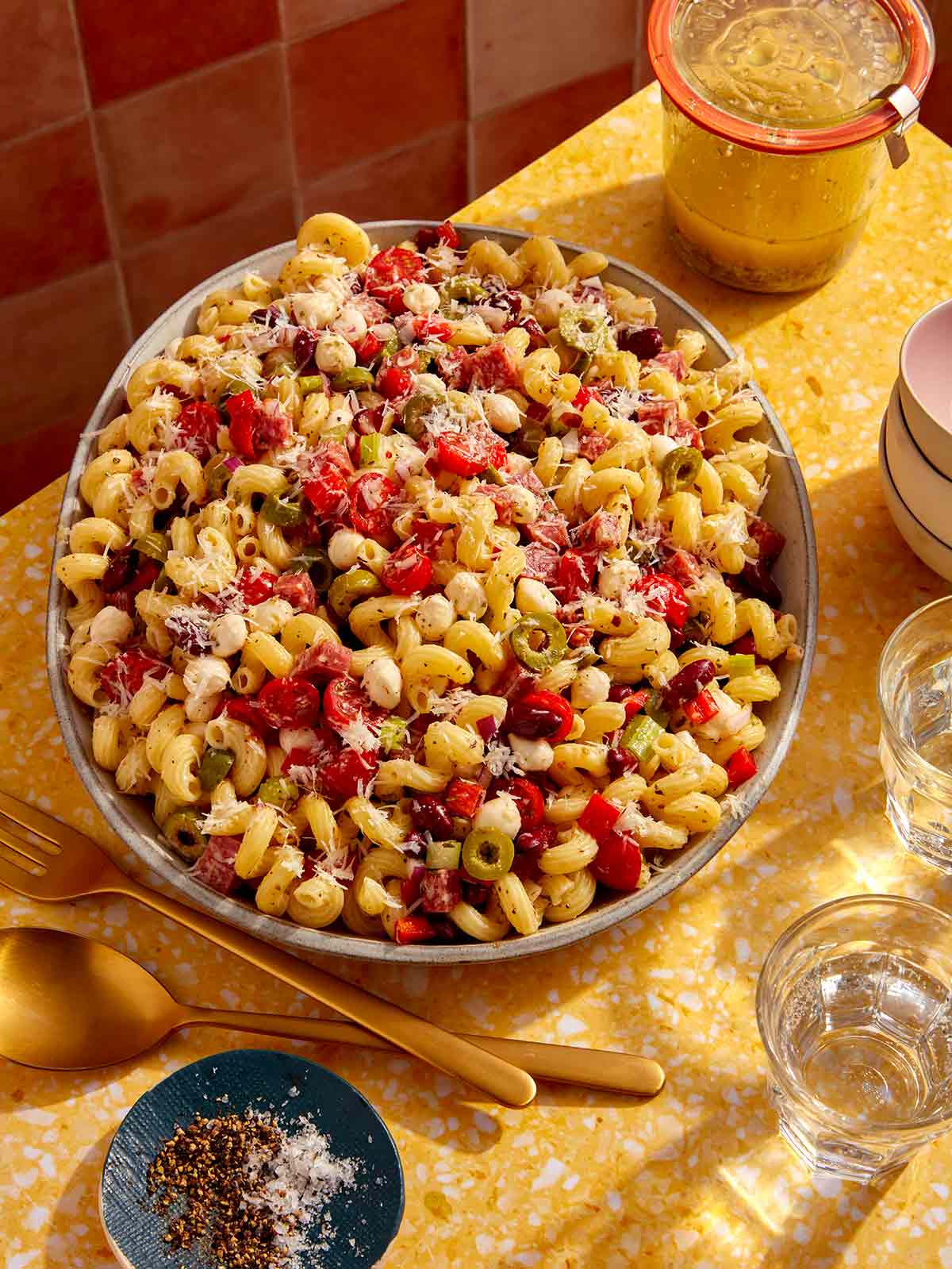 Pasta salad on a table with serving utensils and a jar of Italian vinaigrette.