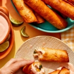 Flautas being served with a hand holding half of one over a plate.
