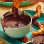 A bowl of homemade chocolate sauce with a churro being dipped in it.
