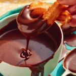 Close up on a bowl of chocolate sauce with a Spanish churro being dipped into it.