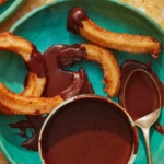 Overhead on a bowl of homemade chocolate sauce with a spoon and churros on the side.