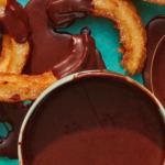 Overhead on a bowl of homemade chocolate sauce with a spoon and churros on the side.